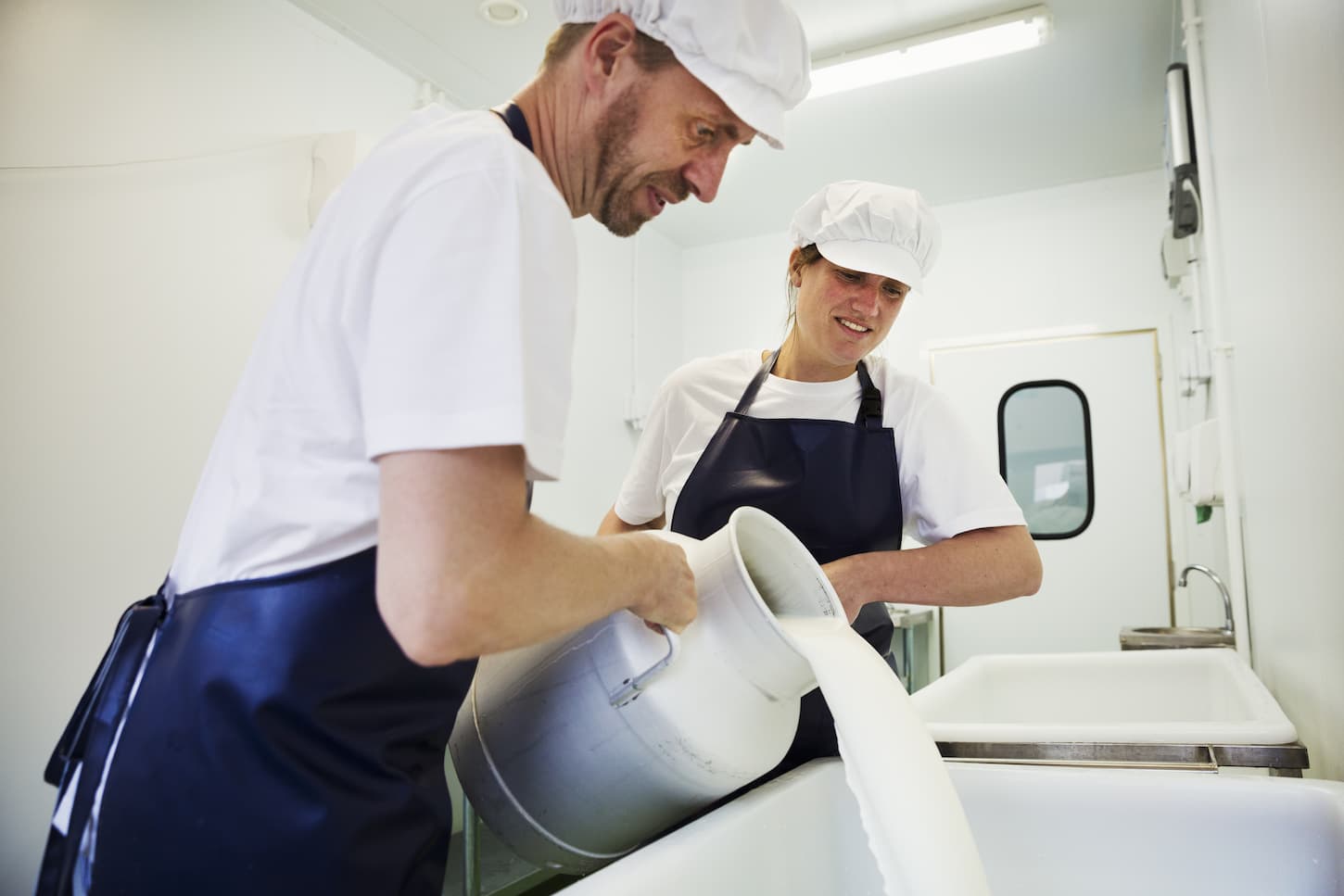 An image of a Man and Woman in a creamery, pouring goat's milk from a churn, making goat cheese.