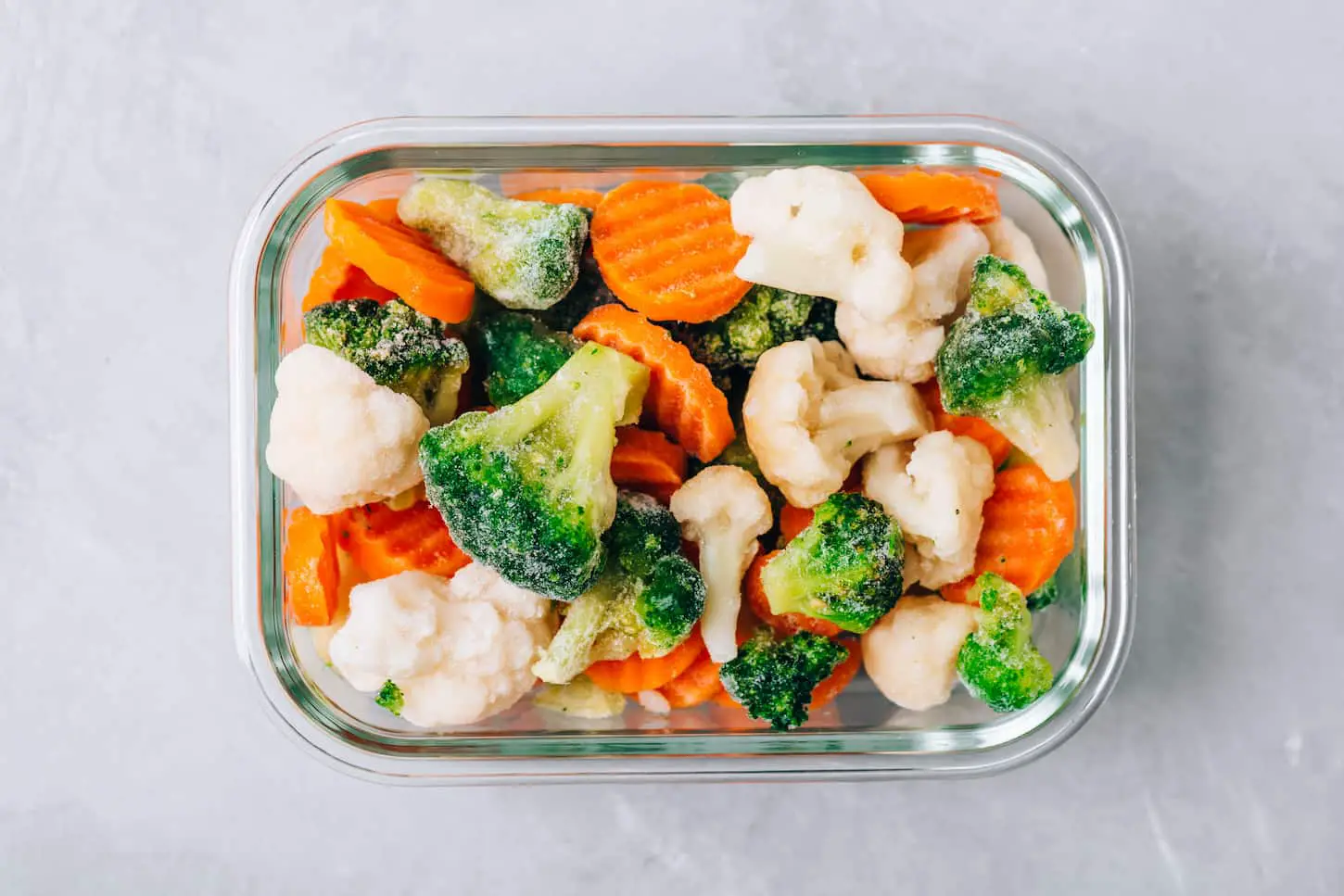 An image of carrots, broccoli, and cauliflower in a glass container on gray stone background.