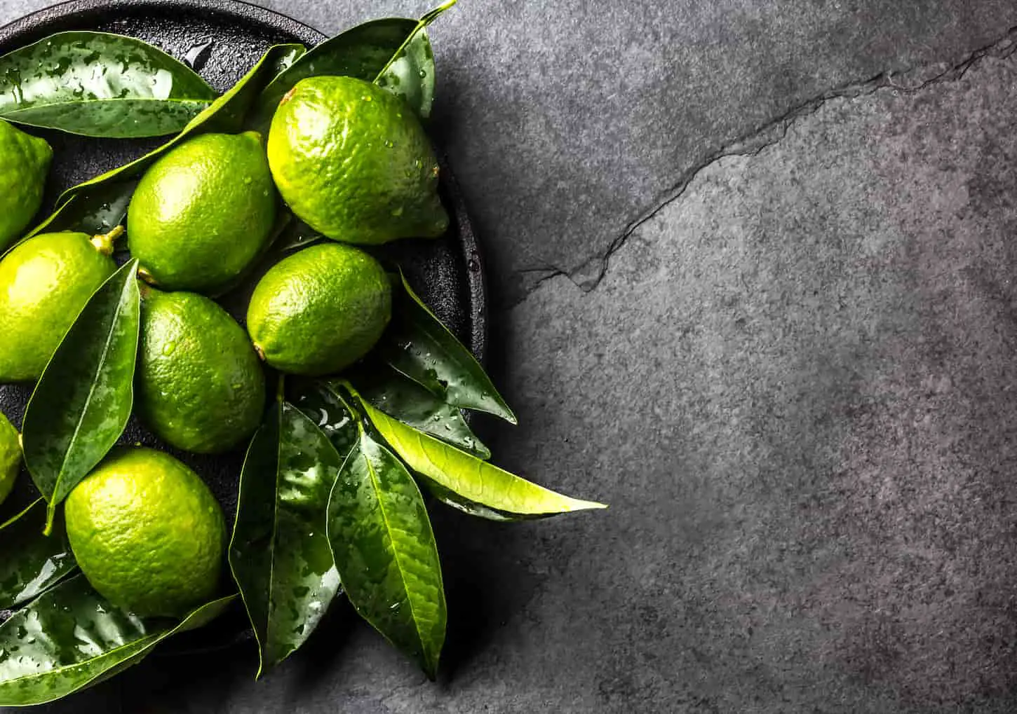 An image of green lemon lime with fresh leaves on a black plate on a dark background.