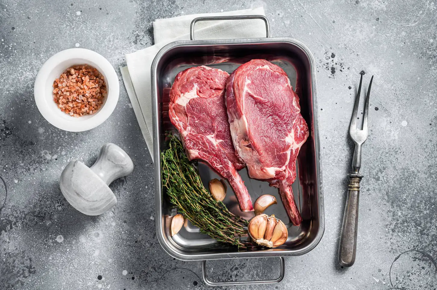 An image of uncooked Raw Tomahawk veal steaks in a steel tray with herbs with a gray background.