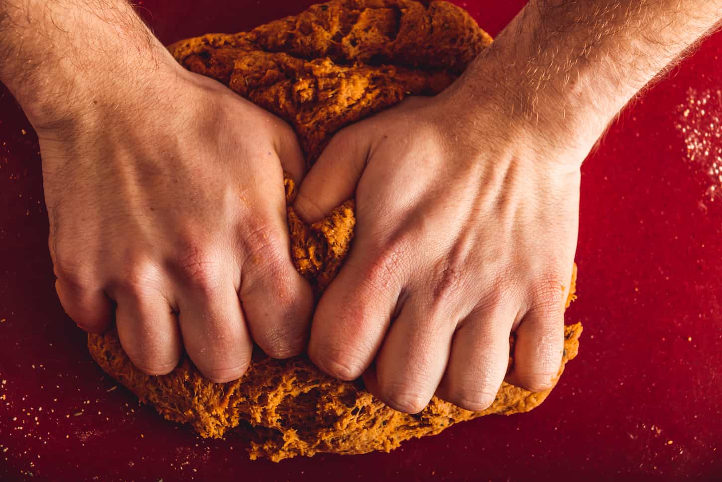 An image focused on a man's hands while kneading seitan dough on a red table.