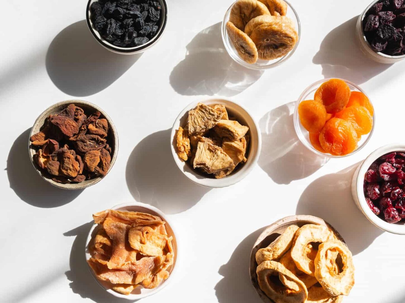 An image of dried or sun-dried berries and fruits in bowls on white background with shadows. 